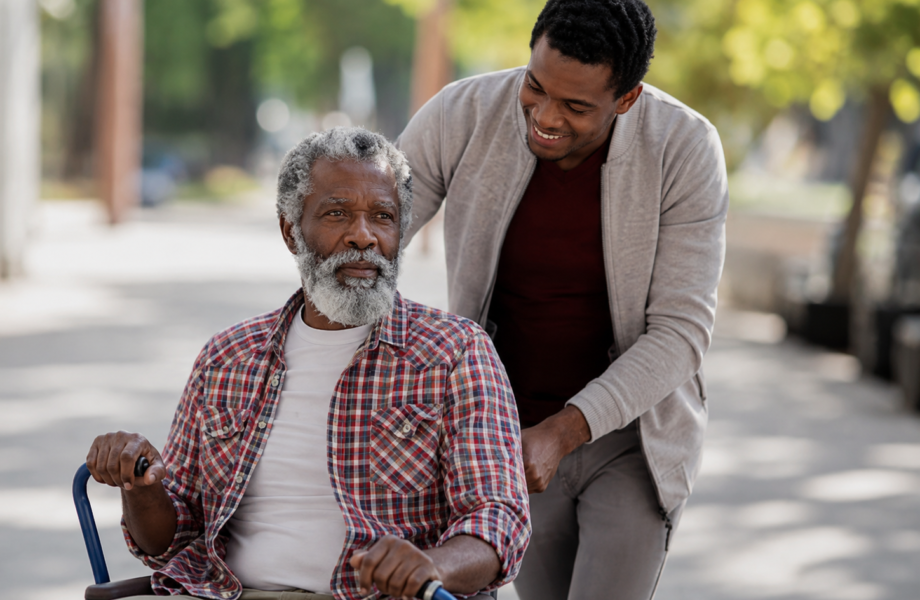 A caregiver assisting an older man in a wheelchair on an outdoor walk